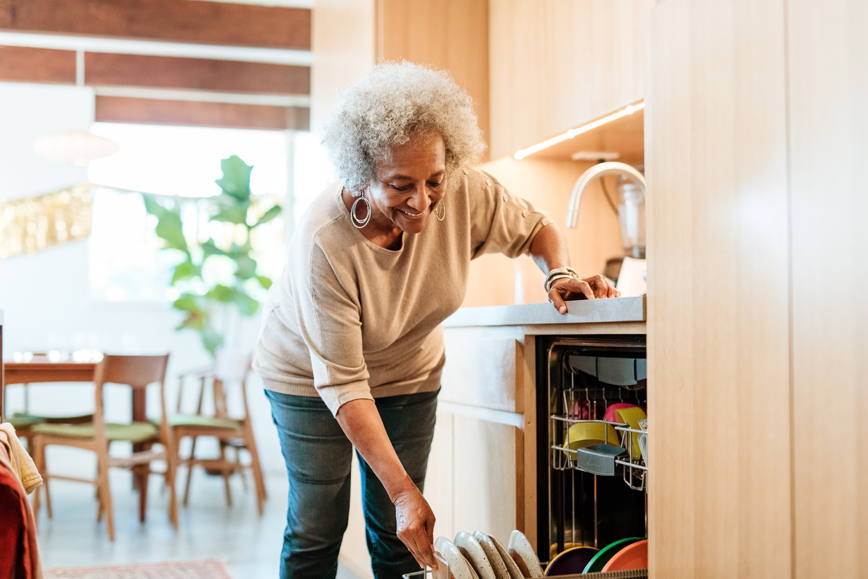 Smiling senior woman keeping plates in dishwasher. Retired elderly female is doing routine chores in kitchen.